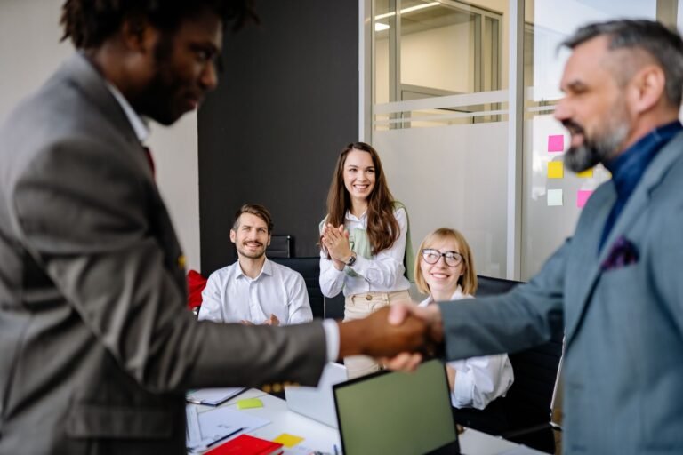 Colleagues in an office cheer as two team members finalize a successful handshake agreement