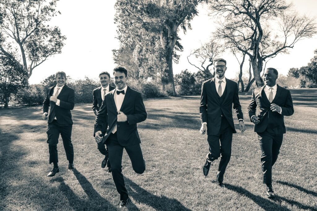Group of groomsmen in suits running across a field candidly