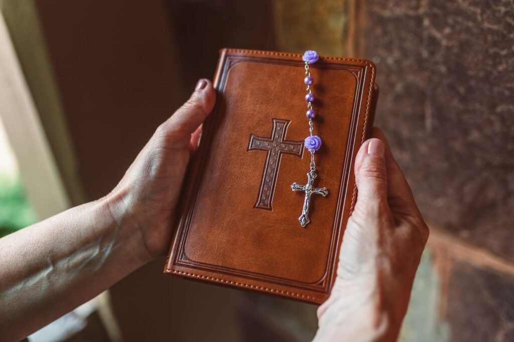 Close-up of hands holding a brown leather Bible with cross and purple rosary beads