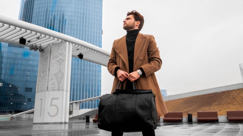 Fashionable man in a coat holding a structured duffel bag near modern architecture