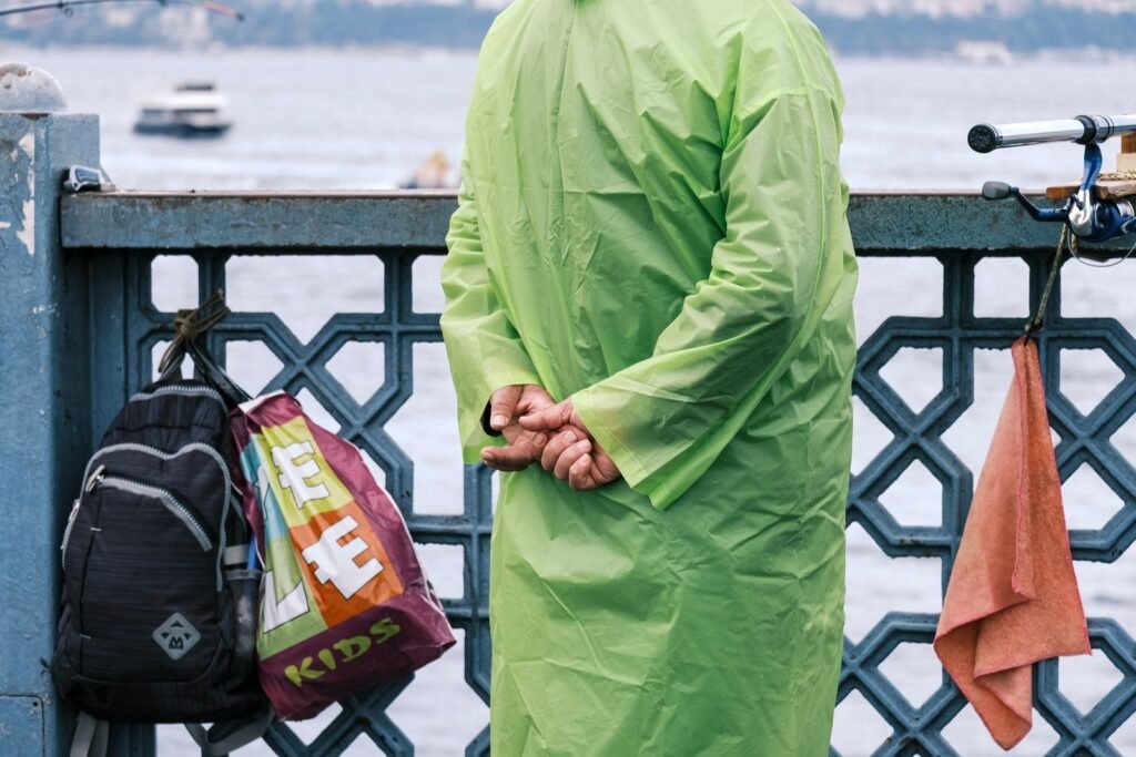 A man in a neon green raincoat stands on a waterfront bridge with bags and a towel