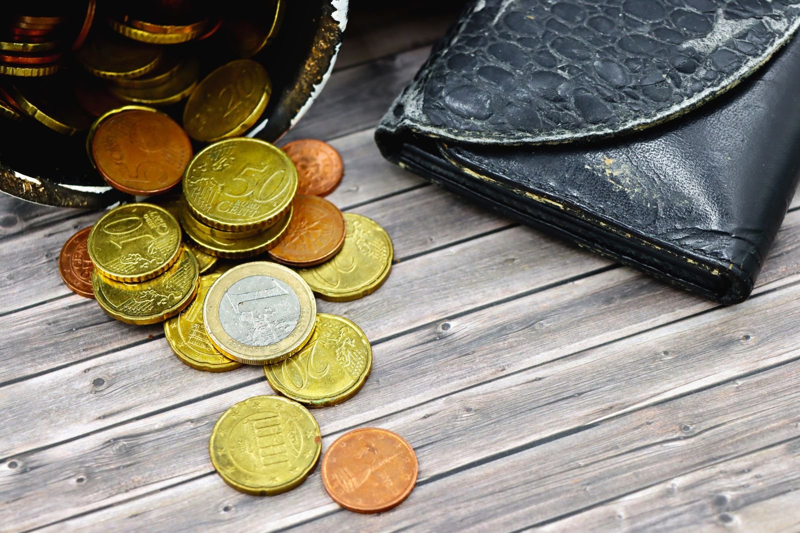 A worn-out leather wallet and scattered euro coins on a wooden surface