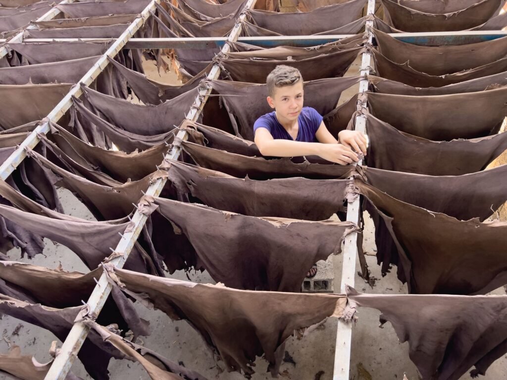 A young boy stands amid drying leather in an outdoor workshop